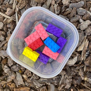 May include: A clear plastic container filled with colorful toy building blocks. The blocks are various colors, including pink, red, yellow, blue, and purple. The container is square with rounded corners.