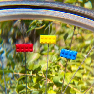 May include: Close-up of a bicycle wheel with three colorful Lego-style brick spoke decorations. The bricks are red, yellow, and blue, attached to the spokes. The background is a blurred view of green foliage.