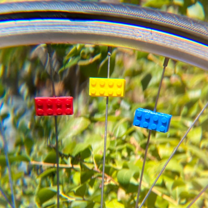 May include: Close-up of a bicycle wheel with three colorful Lego-style brick spoke decorations. The bricks are red, yellow, and blue, attached to the spokes. The background is a blurred view of green foliage.