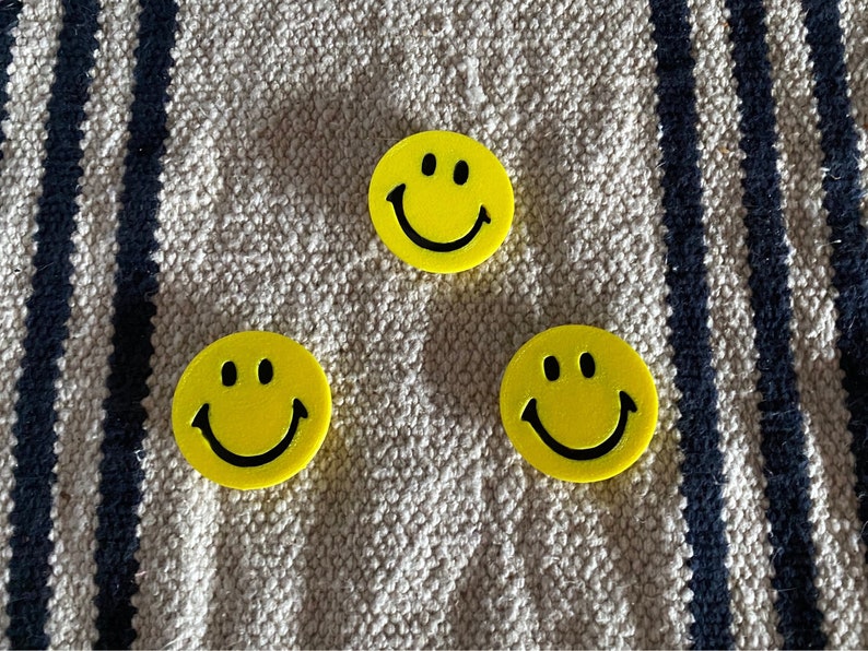 May include: Three yellow smiley face buttons with black eyes and smiles. The buttons are round and have a raised design. They are arranged on a beige and navy blue striped fabric.