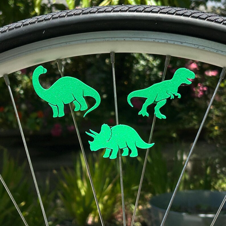 May include: Green dinosaur-shaped bicycle wheel spoke decorations. The image shows three different dinosaur silhouettes: a Brontosaurus, a Tyrannosaurus Rex, and a Triceratops. The decorations are attached to the spokes of a bicycle wheel.