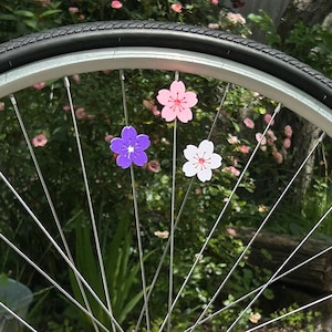 May include: A bicycle wheel with three colorful flower-shaped decorations attached to the spokes. The flowers are pink, white, and purple, with a detailed center. The tire is black, and the background is a blurred green with pink flowers.