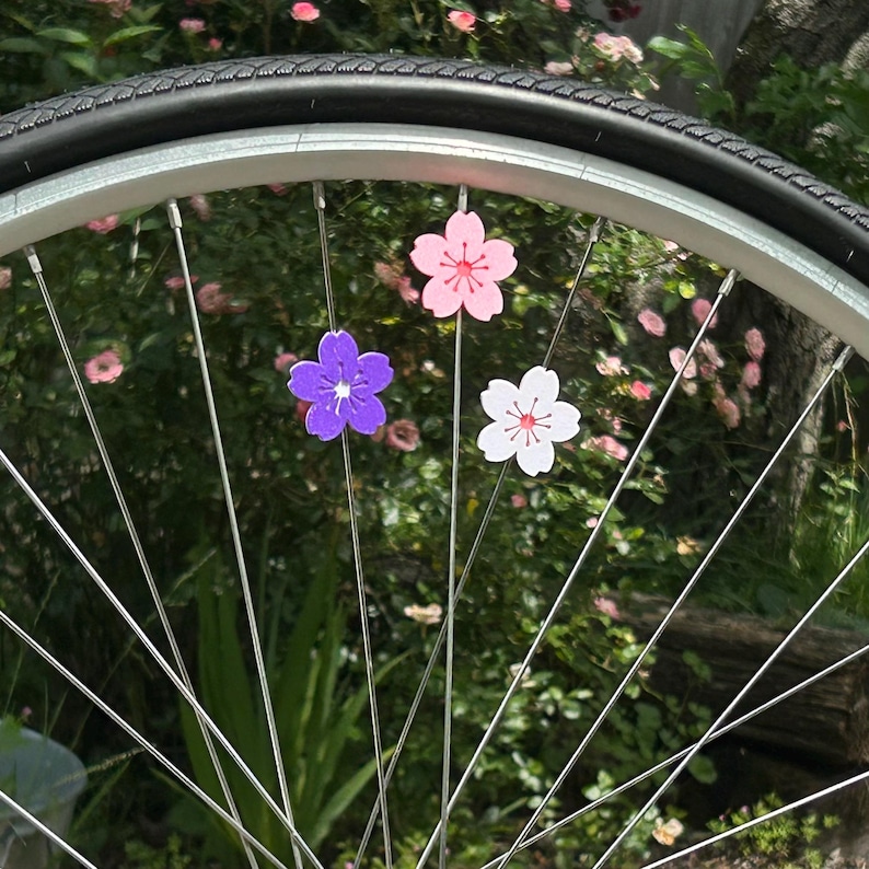 May include: A bicycle wheel with three colorful flower-shaped decorations attached to the spokes. The flowers are pink, white, and purple, with a detailed center. The tire is black, and the background is a blurred green with pink flowers.