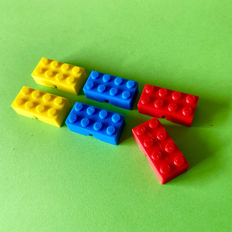 May include: Six colorful toy building blocks in yellow, blue, and red are arranged on a green surface. The blocks are rectangular with circular studs on top, typical of construction toys. The blocks are a classic toy for children.