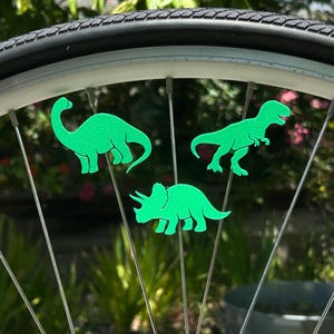 May include: Close-up of a bicycle wheel with three bright green dinosaur-shaped decorations attached to the spokes. The dinosaurs include a Brontosaurus, a Tyrannosaurus Rex, and a Triceratops. The background is blurred green foliage.