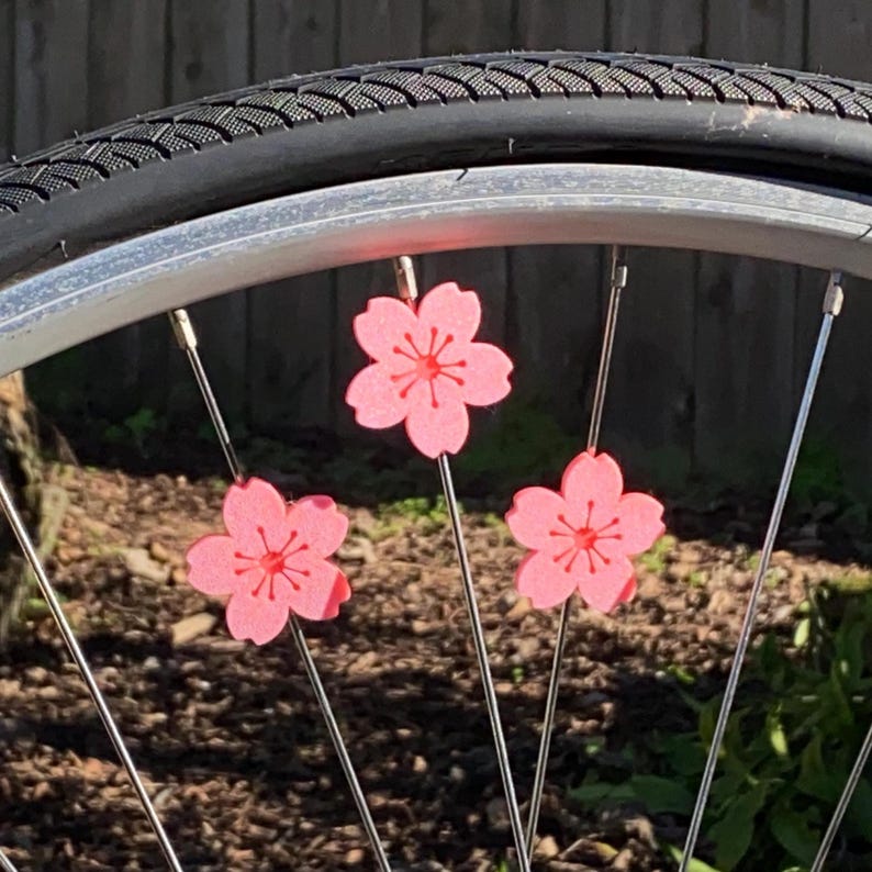 May include: A bicycle wheel with three pink flower-shaped spoke decorations. The flowers have a darker pink center and are attached to the spokes. The tire is black, and the wheel rim is silver.