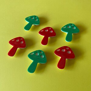 Puede incluir: Una colecci&oacute;n de seis botones con forma de seta dispuestos en un patr&oacute;n disperso. Tres botones son de color rojo vibrante y tres son de color verde brillante. Cada bot&oacute;n presenta una base blanca y tres recortes circulares en la tapa.