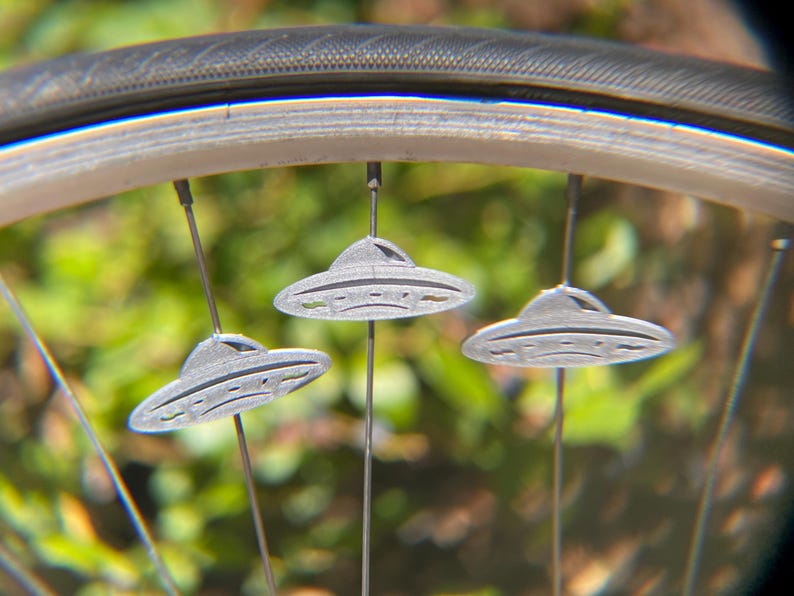 May include: Close-up of a bicycle wheel with three gray, UFO-shaped spoke decorations. The decorations are suspended from the spokes and have a detailed, saucer-like design. The tire is black, and the background is blurred green.