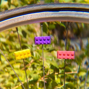 May include: Close-up of a bicycle wheel with colorful, toy brick spoke decorations. The bricks are yellow, purple, and pink, adding a playful touch to the bike's design. The background is a blurred green foliage.