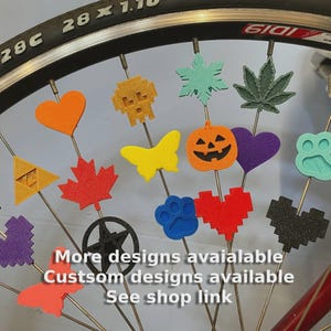 May include: A bicycle wheel with various colorful spoke decorations. The decorations include shapes like hearts, a skull, a maple leaf, a butterfly, a pumpkin, and a paw print. Text on the image says "More designs available, Custom designs available, See shop link."