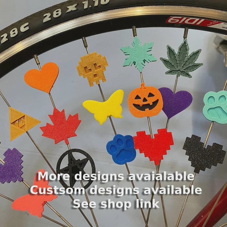 May include: A bicycle wheel with various colorful spoke decorations. The decorations include shapes like hearts, a skull, a maple leaf, a butterfly, a pumpkin, and a paw print. Text on the image says "More designs available, Custom designs available, See shop link."