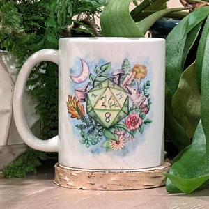 Op de afbeelding: Witte keramische mok met een groene, blauwe en roze illustratie van een 20-zijdige dobbelsteen omringd door bloemen, paddenstoelen en bladeren. De dobbelsteen heeft de nummers 2, 4, 6, 8, 10, 12, 14, 16, 18 en 20.