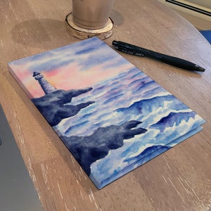 Op de afbeelding: Een aquarel schilderij van een vuurtoren op een rotsachtige kust met blauwe golven die tegen de rotsen breken. De lucht is zacht roze en oranje.