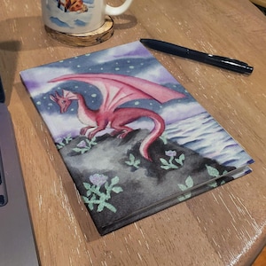 Puede incluir: Un diario de temática fantástica con un dragón rojo de alas rosas en la portada, sobre un fondo de cielo nocturno y mar. La ilustración incluye un dragón detallado sobre un afloramiento rocoso con detalles florales.