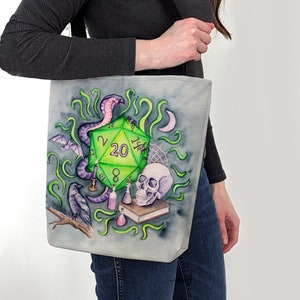 May include: A canvas tote bag featuring a watercolor design with a green twenty-sided die as the central element. The design includes a purple snake, skull, raven, potion bottles, and a bat, all set against a gray background. The bag has a black strap.
