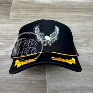 May include: Black trucker hat with a gold trim and a white eagle patch. The eagle is flying with its wings spread. The hat has a mesh back.