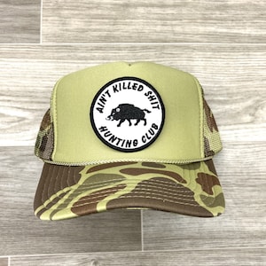 May include: A green and brown camouflage trucker hat with a black and white patch that says "Ain't Killed Shit Hunting Club" and a black silhouette of a wild boar.