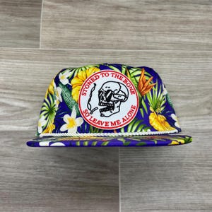 May include: A blue and floral patterned trucker hat with a white rope brim. The hat features a circular patch with a skull smoking a cigarette and the text "STONED TO THE BONE SO LEAVE ME ALONE."