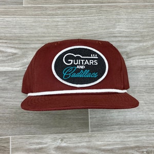 May include: A maroon baseball cap with a white stripe around the brim. The cap has a black and white patch with a guitar and the text "GUITARS AND Cadillacs" in white and teal.