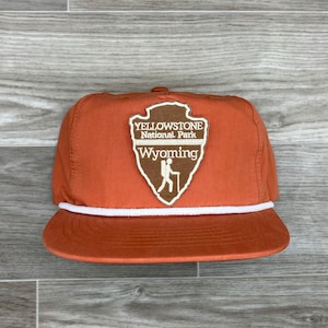 Puede incluir: Una gorra de béisbol vintage de color naranja y blanco con un parche marrón que muestra el logotipo del Parque Nacional de Yellowstone y el texto "Yellowstone National Park Wyoming".
