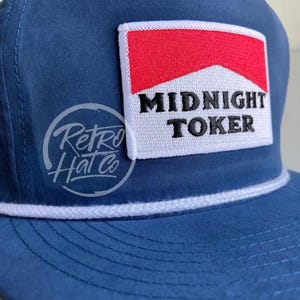 May include: Navy blue trucker hat with a white rope detail and a rectangular patch. The patch features a red and white design with the words "MIDNIGHT TOKER" in black. The hat also has a "Retro Hat Co" logo.