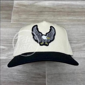 May include: A white and black baseball cap with an embroidered eagle design. The eagle is in flight with its wings spread wide. The cap has a black brim and a white crown.
