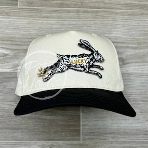 Peut inclure: Casquette de baseball crème avec une visière noire. La casquette présente une illustration en noir et blanc d'un lapin courant avec le mot "LUCKY" en lettres dorées. Une éperon doré fait également partie du design.