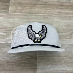 May include: A gray baseball cap with a black brim and a white eagle patch on the front.