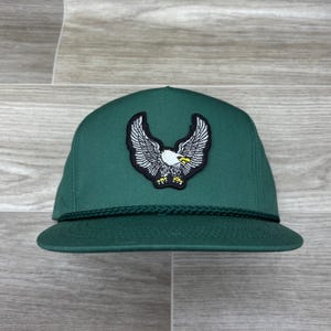 May include: A dark green baseball cap with a black rope trim and an embroidered white eagle with yellow accents.