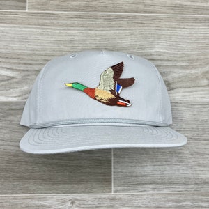 Puede incluir: Una gorra de béisbol gris con un pato silvestre bordado volando en la parte delantera. El pato es marrón, verde y azul.