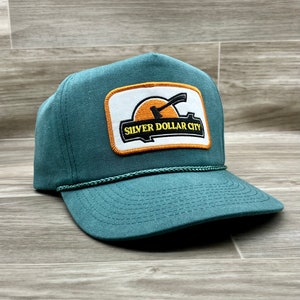 Op de afbeelding: Een turquoise truckercap met een patch waarop "Silver Dollar City" in gele letters staat. De patch toont een zwart silhouet van een zonsondergang met een houweel.