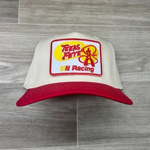 May include: A cream-coloured baseball cap with a red peak. The front displays a rectangular patch with the words "Texas Pete" in yellow and red, a logo, and the word "Racing".