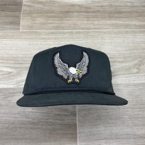 May include: A black baseball cap with a white eagle patch on the front. The eagle has its wings spread and is facing forward. The cap has a black brim and a black strap.