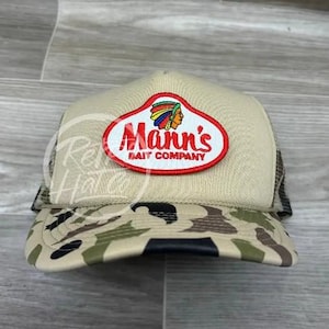 May include: A beige and camouflage trucker hat with a red, white, and blue patch that says "Mann's Bait Company" with an image of an American Indian wearing a feathered headdress.