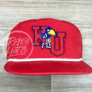 Puede incluir: Gorra de béisbol roja con ribete de cuerda blanca y un parche bordado azul y amarillo que dice "KU" con un pájaro de dibujos animados.