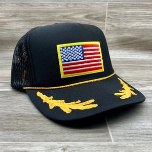 May include: Black trucker hat with a yellow embroidered American flag on the front and gold embroidered details on the brim.