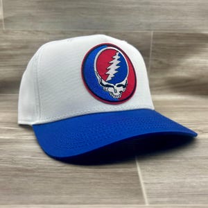 May include: A white baseball cap featuring a circular embroidered patch on the front. The patch displays a blue and red design with a white lightning bolt skull graphic. The cap has a solid blue brim and a structured crown with white stitching.
