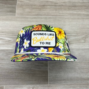 Puede incluir: Una gorra de béisbol de estampado tropical morado y verde con ribete de cuerda blanca y un parche negro que dice "Sounds Like Bullshit To Me".