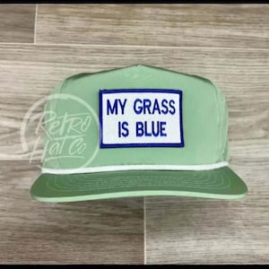 以下が含まれることがあります： 白いつばと「My Grass Is Blue」と書かれた青と白のパッチが付いた薄い緑色のトラッカーハットです。
