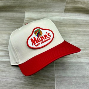 Puede incluir: Una gorra de béisbol blanca y roja con visera roja. La parte delantera de la gorra tiene un parche con un tocado de nativo americano y el texto "Mann's Bait Company."