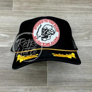 May include: Black trucker hat with a mesh back and a circular patch featuring a skull smoking a cigarette. The patch reads "STONED TO THE BONE, SO LEAVE ME ALONE" in a red and white design. The hat has yellow accents.