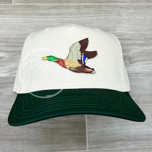 Puede incluir: Una gorra de béisbol blanca con la parte inferior verde. La gorra tiene una imagen bordada de un pato silvestre en vuelo.