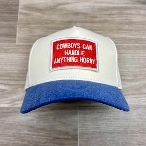 Peut inclure: Casquette de baseball blanche et bleue avec le texte "COWBOYS CAN HANDLE ANYTHING HORNY" dans un rectangle rouge. La casquette a une visière bleue et un panneau avant blanc.