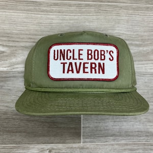 Puede incluir: Un gorro de camionero verde con un parche rojo y blanco que dice "Uncle Bob's Tavern".
