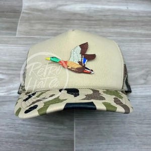 May include: A beige and camouflage trucker hat with a brown, green, and blue embroidered mallard duck flying on the front.