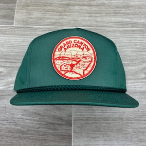 May include: A green baseball cap with a flat brim and a black rope detail. The cap features a circular patch with the words "Grand Canyon Arizona" in red and white, depicting a canyon landscape.