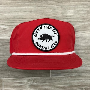 May include: Red baseball cap with a white rope trim and a black and white patch that says "Ain't Killed Shit Hunting Club" with a boar silhouette.