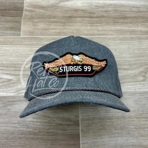 Puede incluir: Una gorra de camionero gris oscuro con un parche que muestra un águila y el texto "Sturgis 99".