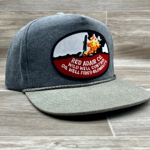 May include: A gray and green trucker hat with a red, white, and yellow patch that reads "Red Adair Co. Oil Wild Well Control Well Fires-Blowouts".