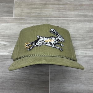May include: Olive green corduroy baseball cap with a running rabbit patch. The patch features a black and white rabbit with the word "LUCKY" in gold. The cap has a curved brim and a braided rope detail.